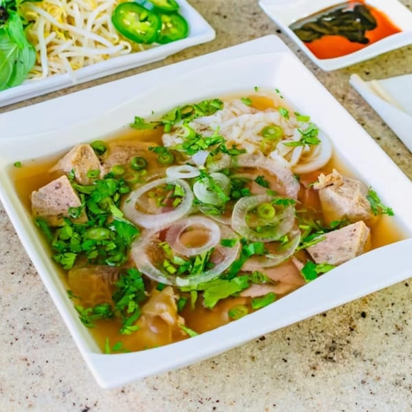 A bowl of vPho's Vietnamese pho with meat, onions, and herbs, with sides of bean sprouts and jalapeños.