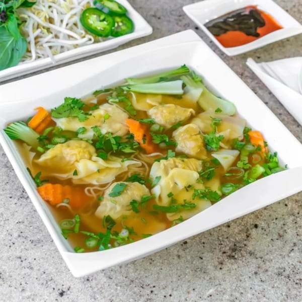 A white bowl of Vietnamese wonton soup with carrots, bok choy, and cilantro, with sides of bean sprouts and jalapeños.