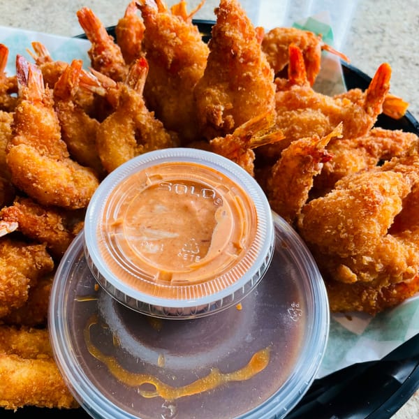 Crispy fried shrimp with tails on, served in a black basket with two containers of dipping sauce.
